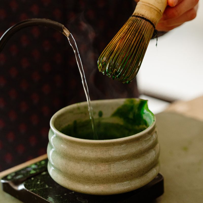 Hot water poured into a bowl of bright green matcha, whisk ready